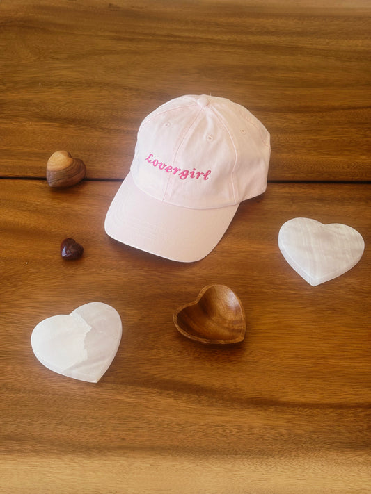 Pink cap with 'Lovergirl' text on a wooden surface with heart-shaped objects.